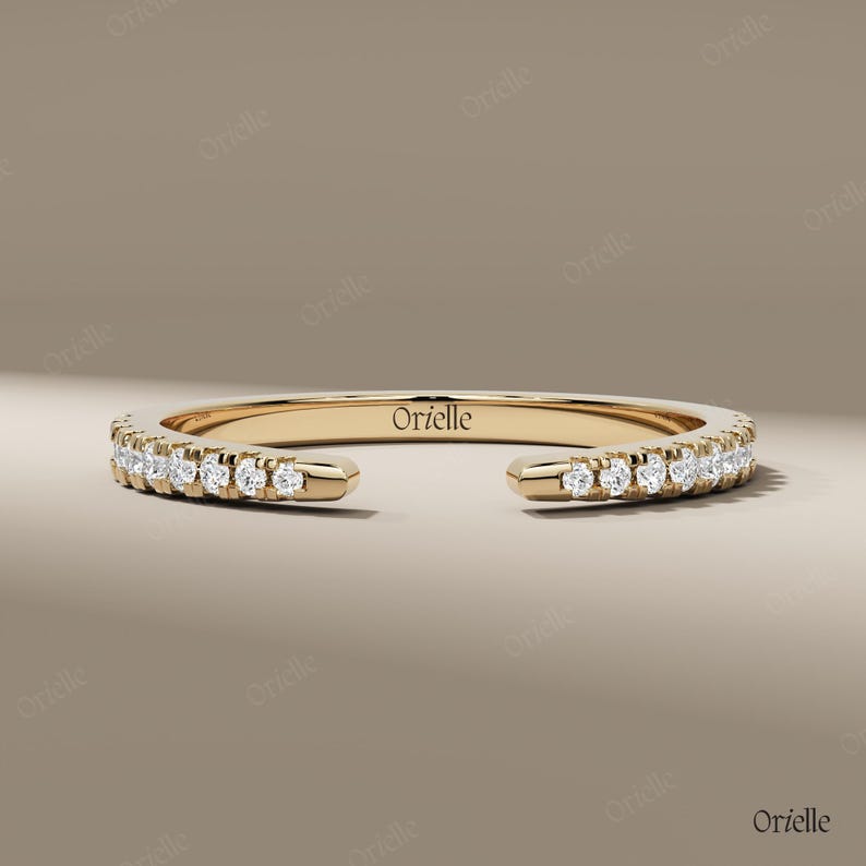 May include: A gold-colored ring with a row of small, clear gemstones set along the top half. The ring has an open design, with the ends not connected. The word "Orielle" is engraved on the inside of the ring.