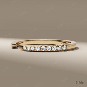 May include: A gold-colored ring with a row of small, clear gemstones set along the top. The ring has a modern, open design, with one end curving away from the main band. The gemstones sparkle against the gold.