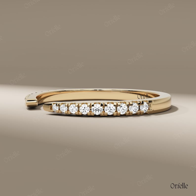 May include: A gold-colored ring with a row of small, clear gemstones set along the top. The ring has a modern, open design, with one end curving away from the main band. The gemstones sparkle against the gold.
