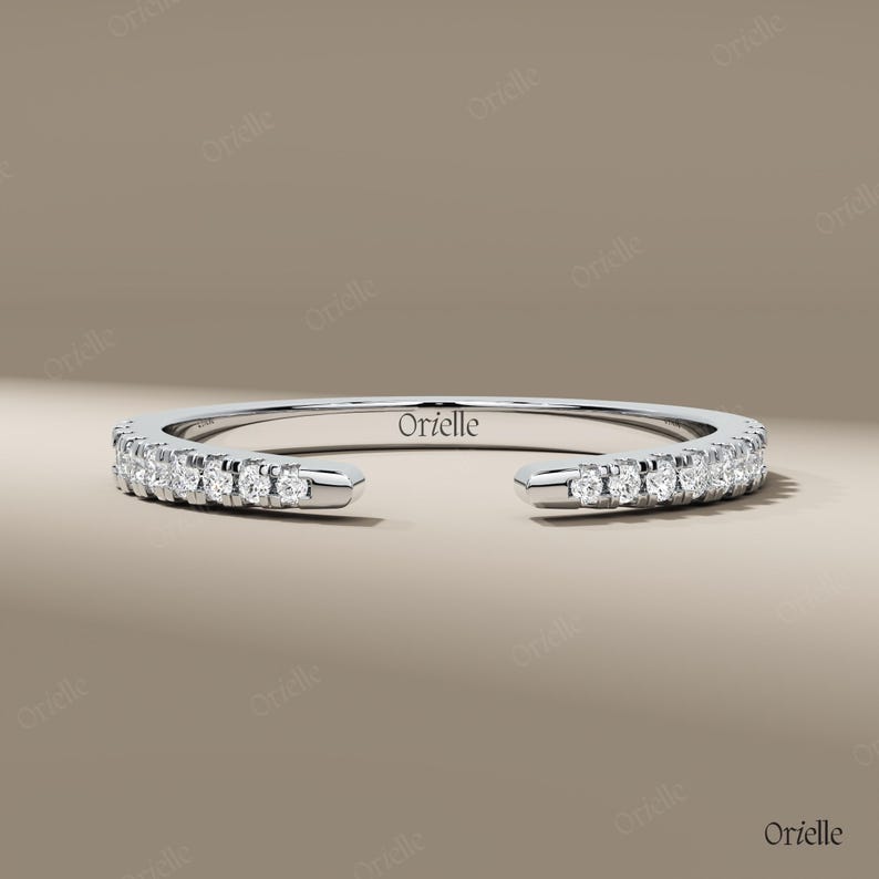 May include: A silver-colored ring with a row of small, clear gemstones. The ring has an open design, with the ends of the band not connected. The word "Orielle" is engraved on the ring. The ring is displayed on a neutral background.