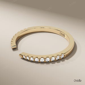 May include: A gold-colored ring with a row of small, clear gemstones set along the front. The ring has an open design, with a gap at the front. The word "Orielle" is engraved on the ring.