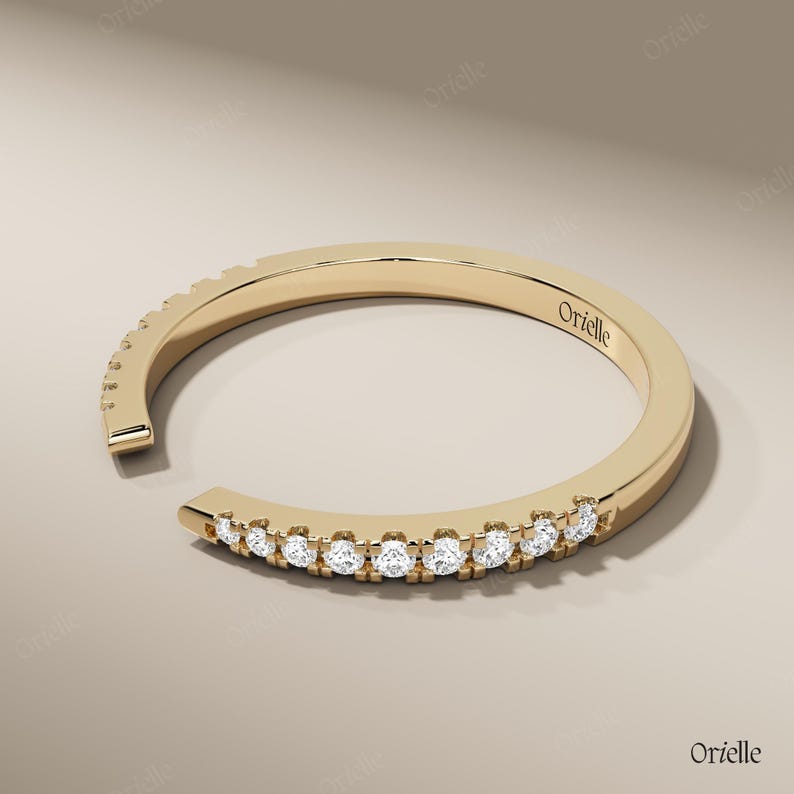 May include: A gold-colored ring with a row of small, clear gemstones set along the front. The ring has an open design, with a gap at the front. The word "Orielle" is engraved on the ring.