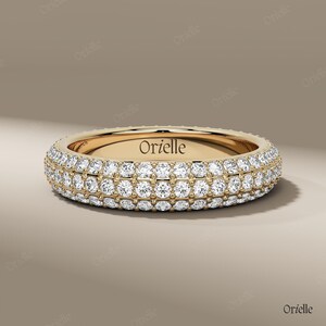Puede incluir: Un anillo de oro con tres filas de brillantes gemas blancas. El anillo tiene forma redondeada y el nombre de la marca "Orielle" está grabado en el interior. El anillo está sobre un fondo neutro.