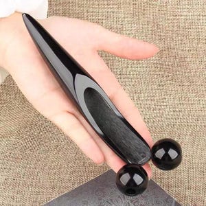 May include: A black massage tool with a long, curved handle and two round balls. The tool is made of a shiny, smooth material and is designed for body massage. The balls are approximately 1 inch in diameter.