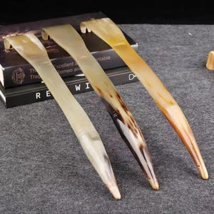 Puede incluir: Tres calzadores con forma de cuerno en tonos beige y marrón. Los calzadores tienen un mango curvo y una punta. Están colocados sobre una superficie gris, con libros en el fondo.