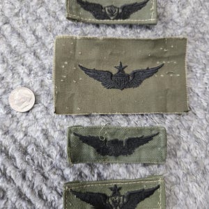 May include: Four olive green fabric patches with black embroidered wing designs. The patches vary in size and shape, with one larger rectangular patch and three smaller rectangular patches. A US dime is next to the patches.