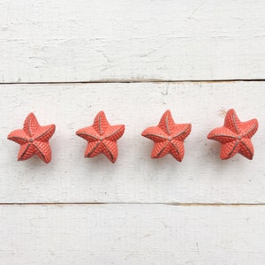 May include: Four coral-colored starfish arranged in a row on a white wooden surface.