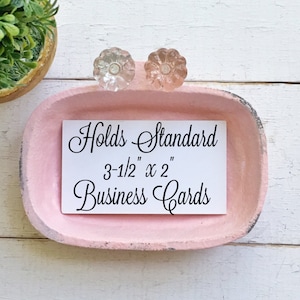 May include: A pink ceramic dish with two glass knobs on top. The dish is designed to hold standard 3-1/2" x 2" business cards. The text "Holds Standard 3-1/2" x 2" Business Cards" is printed on a white card inside the dish.