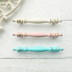 May include: Three decorative cabinet pulls in cream, pink, and turquoise, each with gold accents. The pulls are arranged horizontally on a white wooden surface. The design is vintage-inspired, with a distressed finish.