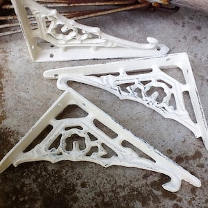 May include: Three white cast iron shelf brackets with a decorative floral design. The brackets are in a distressed finish, giving them a vintage look.