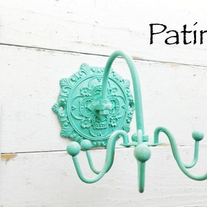 May include: A turquoise metal wall hook with a decorative floral design and three hooks. The word "Patina" is written in black above the hook.