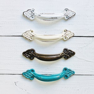 May include: Four decorative cabinet pulls in distressed finishes: white, cream, bronze, and turquoise. Each pull has an arched handle and ornate end details, perfect for adding a vintage touch to furniture or cabinetry.