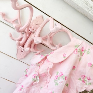 May include: A pink metal coat rack with multiple hooks, hanging on a white wall. A pink dress with a floral pattern hangs on the rack.