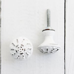 May include: Two white ceramic cabinet knobs with a floral design. The knobs have a distressed finish and are perfect for adding a touch of farmhouse charm to any room.