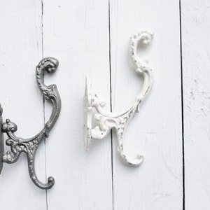 May include: Two ornate metal hooks, one in a silver finish and one in a white finish, mounted on a white wooden surface.