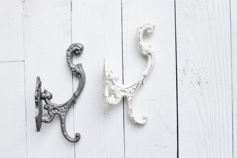 May include: Two ornate metal hooks, one in a silver finish and one in a white finish, mounted on a white wooden surface.