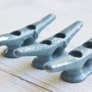 May include: Three blue metal cleat hooks, also known as mooring cleats, are arranged in a row on a white wooden surface. The cleats are designed to secure ropes or lines.