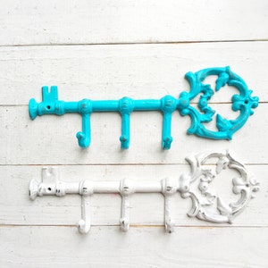 May include: Two decorative key-shaped wall hooks. One is turquoise, the other is white. Each key has three hooks for hanging items. The keys are made of metal and have an ornate, vintage design. The background is a white, distressed wood.