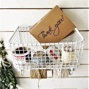 May include: White wire basket with hooks hanging on a white wall. The basket contains several spools of twine and a stack of brown cards with the text "Thank you!" written in black.