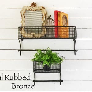 May include: Two black metal wire shelves with a towel bar underneath each shelf. The top shelf has a decorative frame, a birdcage, and a book. The bottom shelf has a potted plant. The shelves are labeled "Oil Rubbed Bronze".