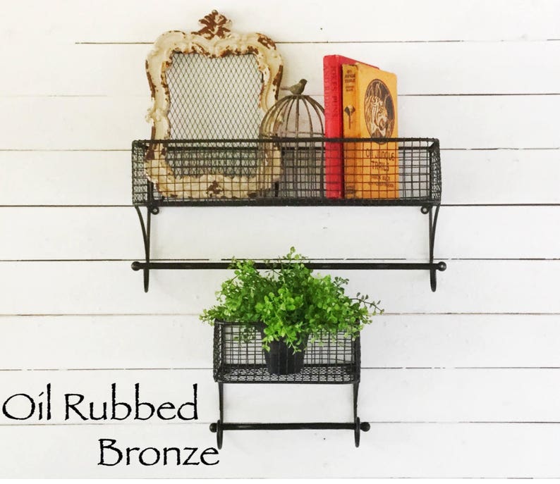 May include: Two black metal wire shelves with a towel bar underneath each shelf. The top shelf has a decorative frame, a birdcage, and a book. The bottom shelf has a potted plant. The shelves are labeled "Oil Rubbed Bronze".