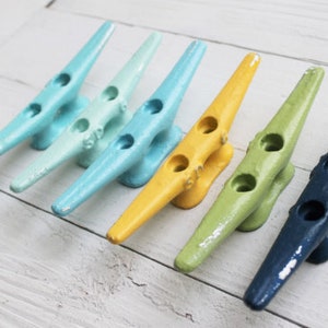 May include: Six colorful boat cleats in a rainbow gradient of blue, green, yellow, and navy blue. The cleats are made of metal and have two holes each.
