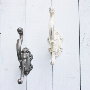 May include: Two decorative wall hooks with an ornate design. One hook is painted white and the other is a dark gray metal.