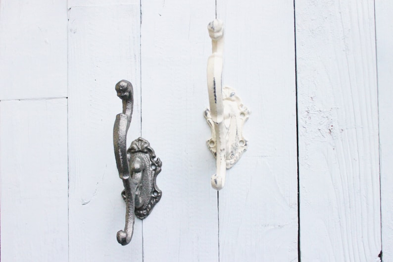 May include: Two decorative wall hooks with an ornate design. One hook is painted white and the other is a dark gray metal.