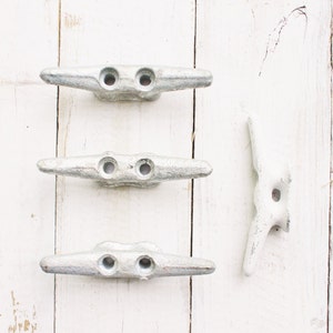 May include: Three silver metal cleat hooks on a white wooden background. The cleat hooks are commonly used for securing ropes on boats.