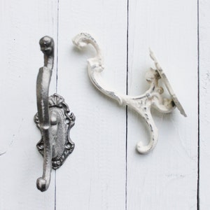 May include: Two decorative wall hooks, one in black and one in white, with ornate designs. The hooks are mounted on a white wooden surface.