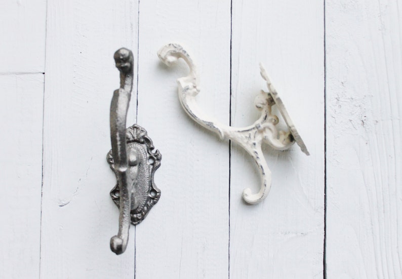 May include: Two decorative wall hooks, one in black and one in white, with ornate designs. The hooks are mounted on a white wooden surface.