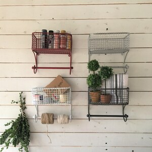 May include: Three sets of wall mounted wire baskets with towel bars. The top baskets are red, silver, and white. The bottom baskets are white, silver, and black. The baskets are filled with various items, including spray paint cans, plants, towels, and twine.