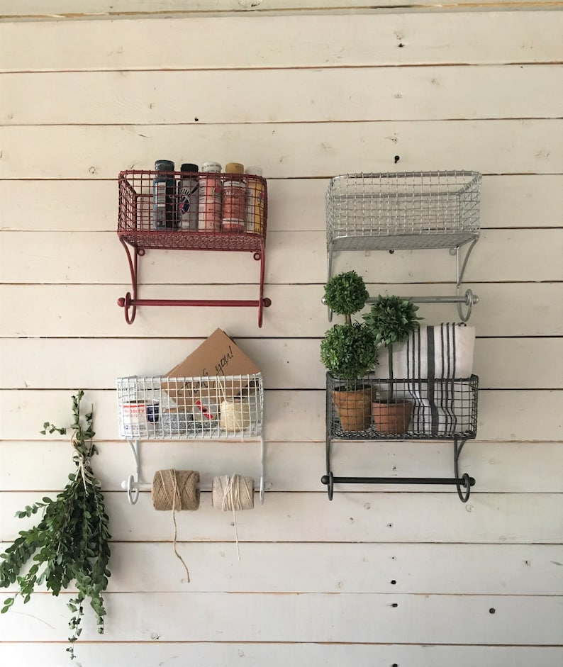 May include: Three sets of wall mounted wire baskets with towel bars. The top baskets are red, silver, and white. The bottom baskets are white, silver, and black. The baskets are filled with various items, including spray paint cans, plants, towels, and twine.