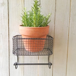 May include: A small potted rosemary plant sits on a black metal shelf with a towel rack underneath. The shelf is mounted to a white wooden wall.