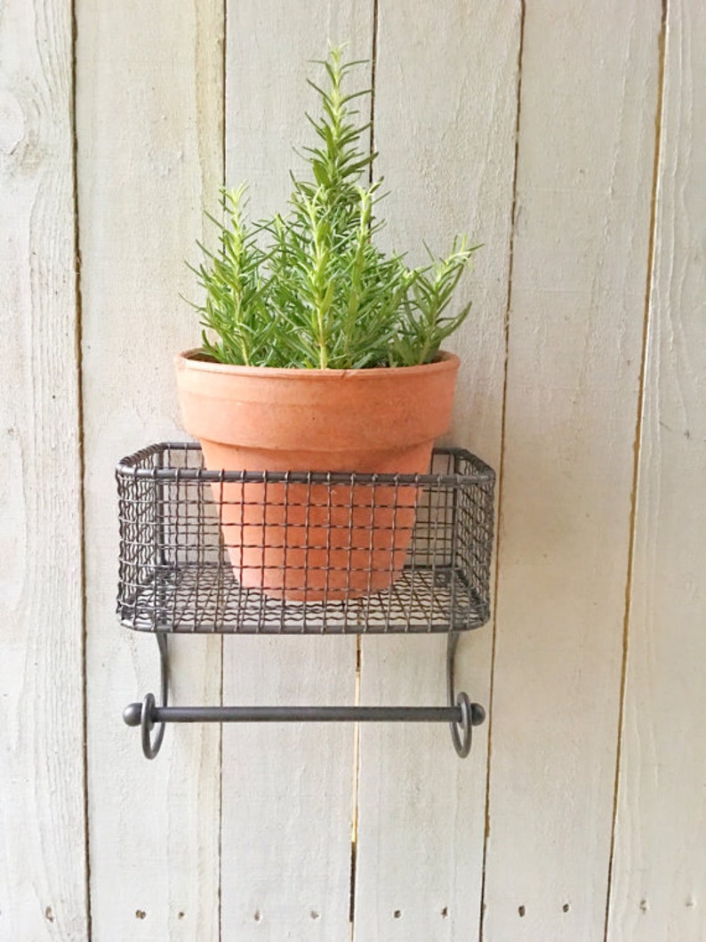 May include: A small potted rosemary plant sits on a black metal shelf with a towel rack underneath. The shelf is mounted to a white wooden wall.