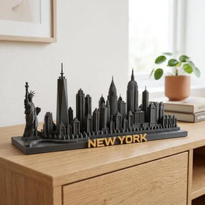 Peut inclure: Un modèle 3D noir mat de la ligne d'horizon de New York représentant la Statue de la Liberté à gauche et divers gratte-ciel. La base est rectangulaire et présente le texte NEW YORK en lettres capitales de couleur dorée sur le bord avant.