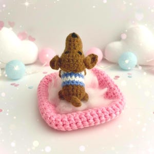May include: A crocheted brown dachshund dog figurine wearing a blue and white striped sweater, resting on a pink heart-shaped bed with white fluff. The background includes decorative elements like heart-shaped pillows and pastel-colored spheres.