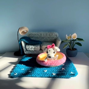 Op de afbeelding: Een gehaakte miniatuurscène met een Dalmatische hond in een roze en geel bed op een blauw tapijt. Een grijze bank, een lamp en een plant in een pot maken de setting compleet. De hond draagt een roze strik.