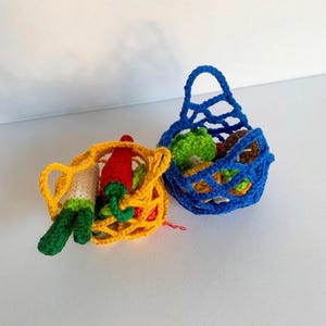 May include: Two miniature crochet market bags, one yellow and one blue, filled with small crocheted vegetables. The yellow bag contains a white and green leek and a red pepper, while the blue bag holds various green and brown crocheted items.