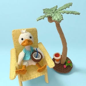 May include: Crocheted duck wearing a blue vest, sitting in a yellow beach chair, holding a coconut drink. A crocheted palm tree and drink are nearby. The scene is set against a light blue background.