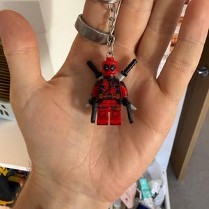 Może przedstawiać: Brelok Lego Deadpool. Figurka jest czerwono-czarna, z dwoma mieczami na plecach i dwoma pistoletami. Brelok ma srebrny pierścień i łańcuszek.
