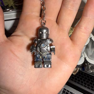 May include: A silver-colored Iron Man Lego figure keychain. The figure has a detailed armor design with a blue circular chest piece and is attached to a silver chain. The figure is approximately 2 inches tall.