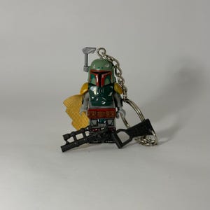 Könnte beinhalten: Ein Boba Fett Lego-Schlüsselanhänger. Die Figur hat einen grünen Helm mit roten Akzenten, einen gelben Umhang und einen schwarzen Blaster. Der Schlüsselanhänger ist an einer silbernen Kette befestigt.