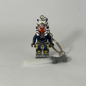 Könnte beinhalten: Ein Star Wars Ahsoka Tano Minifigur-Schlüsselanhänger. Die Figur hat eine weiß-blaue Kopfbedeckung, ein orangefarbenes Gesicht und eine blau-gelbe Rüstung. Ein silberner Schlüsselring und ein weiß-graues Lichtschwert sind befestigt.