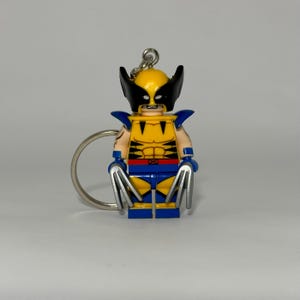 May include: A Wolverine-themed keyring featuring a yellow and blue minifigure with black mask and silver claws. The keyring has a silver ring for attaching to keys or bags. The figure is a stylized representation of the Marvel Comics character.
