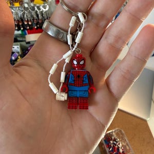 Op de afbeelding: Een sleutelhanger met een rode, blauwe en zwarte Spider-Man Lego-figuur. De figuur is bevestigd aan een zilveren sleutelhanger met een wit koord en witte cilindrische kralen. De figuur is een miniatuurversie van de superheld.