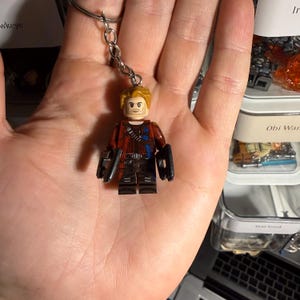 Puede incluir: Un llavero con una minifigura Lego de Star-Lord. La figura tiene una cabeza bronceada con una expresión severa, cabello rubio y una chaqueta roja y negra. El llavero está sujeto a una cadena plateada.
