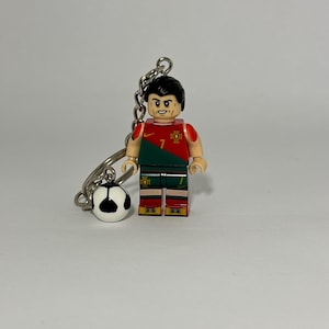 May include: A keychain featuring a Lego-style figurine of a soccer player in a red and green uniform with the number 7. The figurine has a black hairstyle and is attached to a silver chain with a small soccer ball charm.