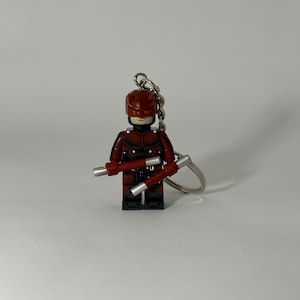May include: A red and black Daredevil Lego keyring. The figure has a red helmet and costume with silver batons. The keyring is attached to a silver chain and ring.