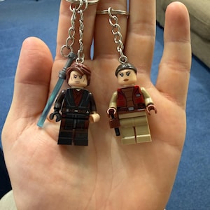 Puede incluir: Dos llaveros estilo Lego con figuras en miniatura. Una figura tiene cabello castaño y un sable de luz, la otra tiene cabello castaño y un chaleco rojo. Ambas figuras están unidas a llaveros plateados.